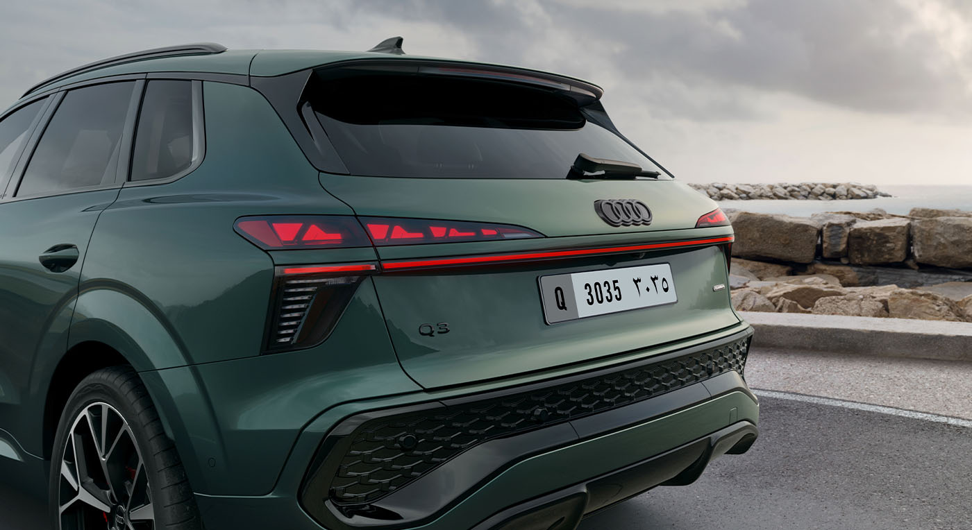 A sleek green Audi Q3 SUV is parked near a rocky shoreline, featuring modern taillights and a rear license plate. The sky is cloudy, suggesting a calm, overcast day.